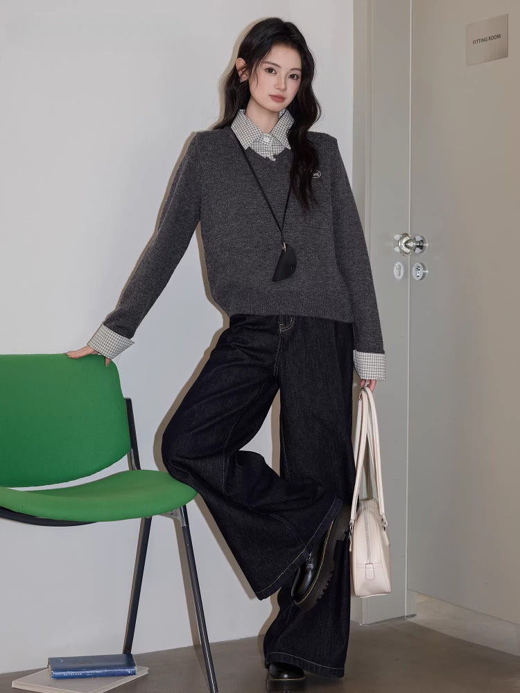 Full body shot of a woman standing with one leg bent, leaning on a green chair, wearing a grey v-neck sweater over a checkered collared shirt, dark wash wide leg jeans, a black crossbody pouch, and holding a white handbag.