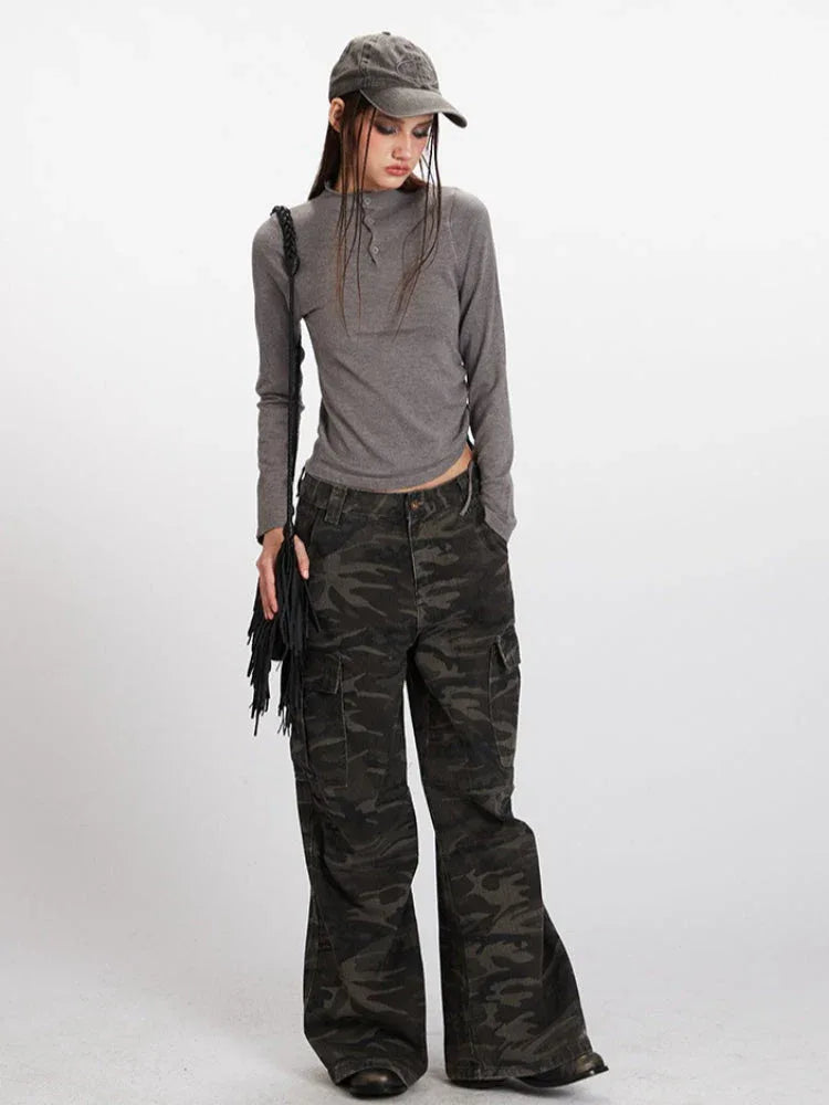 Woman wearing a grey long-sleeved mock neck top with a cropped hem and camouflage print wide-leg cargo pants, accessorized with a grey baseball cap and a black fringe crossbody bag, full body view.