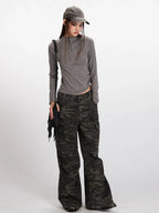 Woman wearing a grey long-sleeved mock neck top with a cropped hem and camouflage print wide-leg cargo pants, accessorized with a grey baseball cap and a black fringe crossbody bag, full body view.