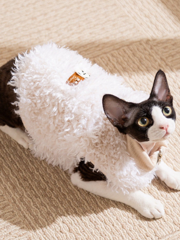 Black and white cat lying down wearing a white sherpa fleece vest with a decorative patch.