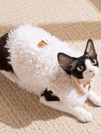 Black and white cat lying down wearing a white sherpa fleece vest with a decorative patch.