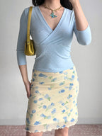 Full body shot of a person wearing a light blue wrap top, a yellow and blue floral skirt, and a gold handbag.