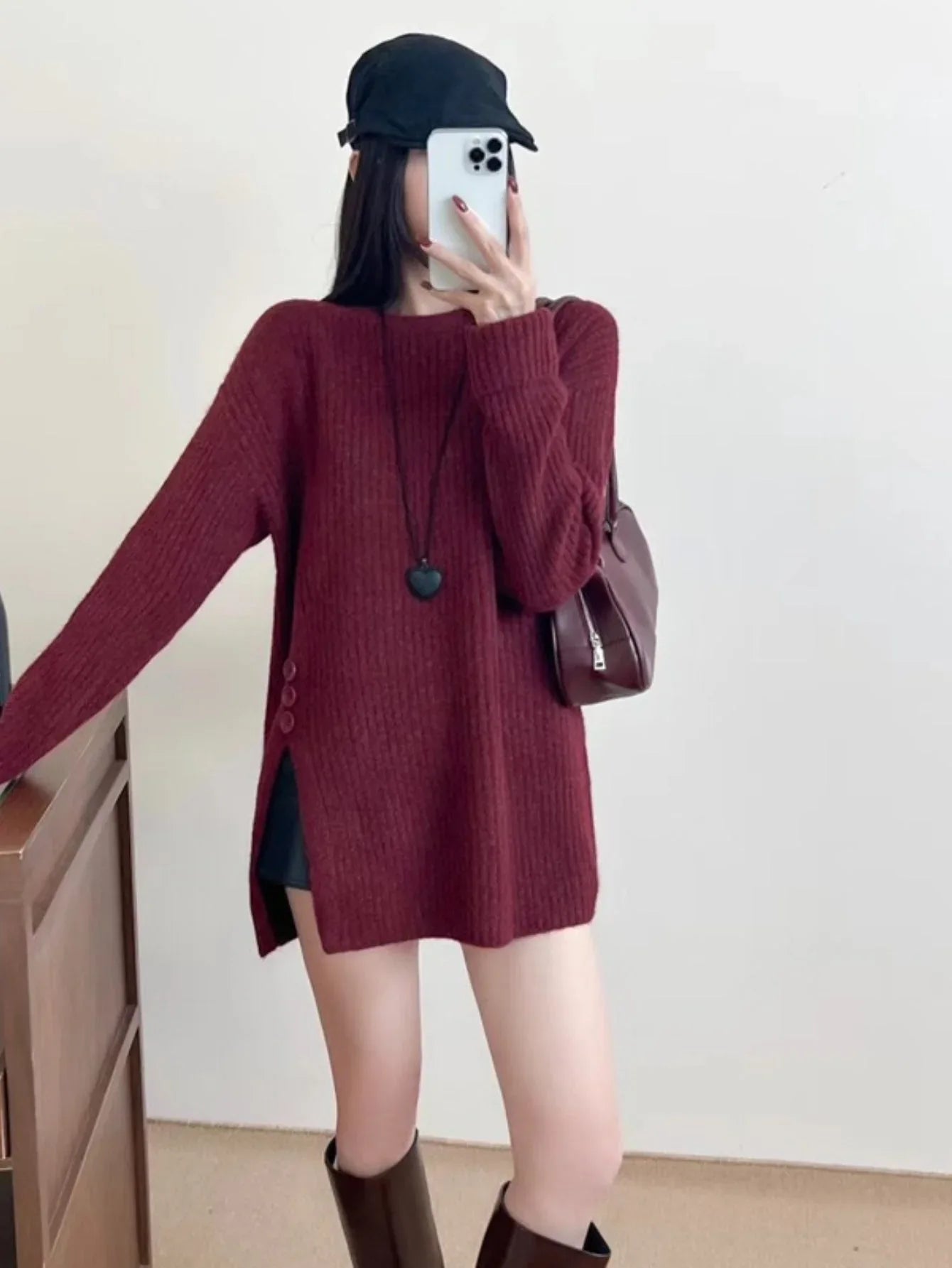 Woman wearing a burgundy ribbed knit longline tunic with side slits and buttons, black shorts, and brown boots, holding a phone to take a mirror selfie.