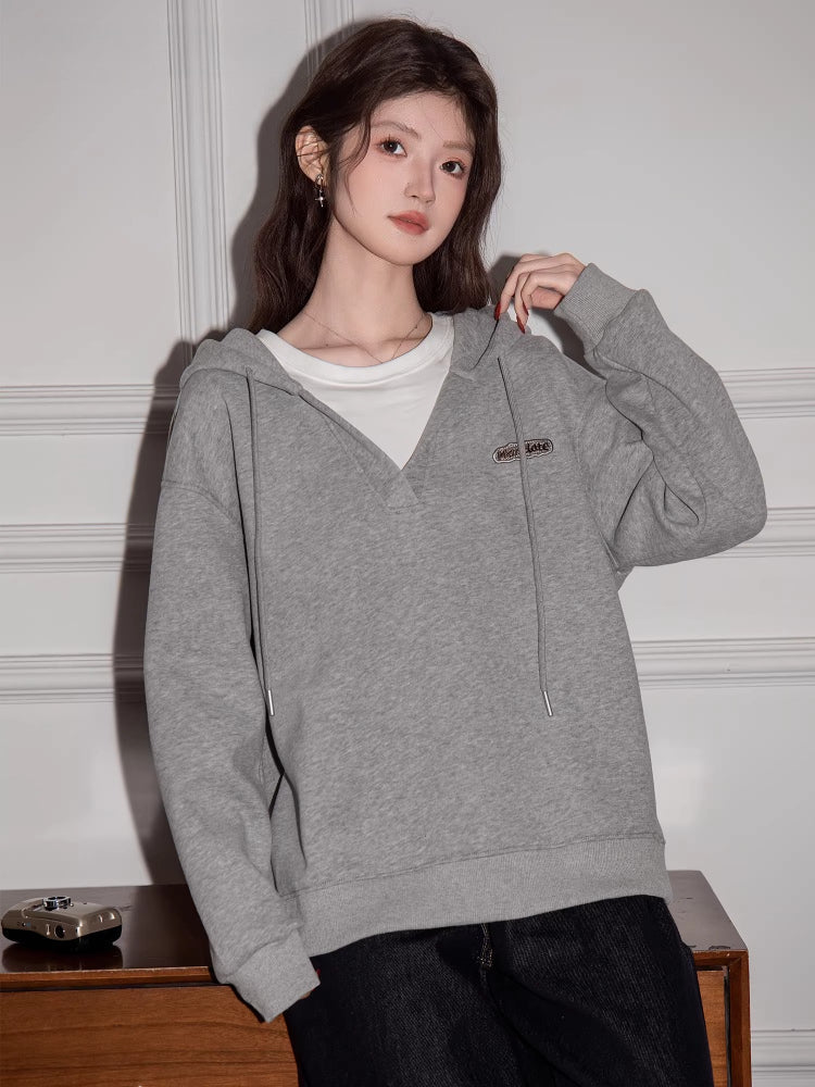 Woman wearing a grey V-neck oversized hoodie over a white t-shirt, standing in front of a white wall.