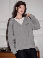 Woman wearing a grey V-neck oversized hoodie over a white t-shirt, standing in front of a white wall.