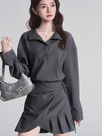 Model holding a silver handbag and wearing a grey long-sleeved collared shirt and a grey skirt.