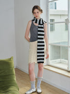 Woman wearing a grey tank top, cream cable knit skirt with fringe, and a black and white striped scarf.