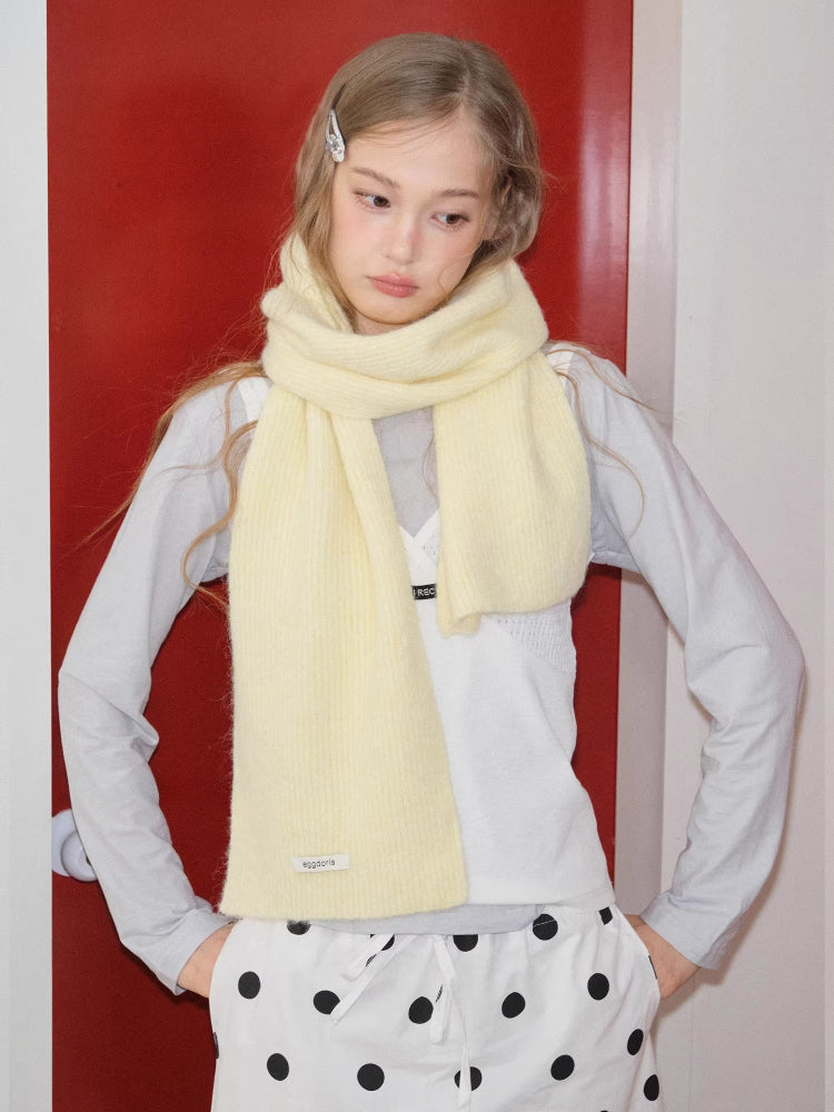 Model wearing a light yellow ribbed knit scarf draped around their neck, paired with a light grey long-sleeved shirt and white polka dot pants.