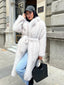 Woman wearing a long, white faux fur coat with a belted waist and blue jeans, holding a black tote bag, outdoors.