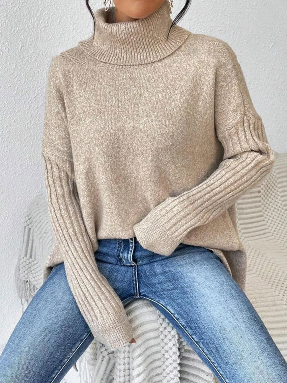 Woman sitting down wearing a beige oversized turtleneck sweater with an asymmetrical hem and ribbed sleeves, paired with blue jeans.