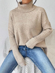 Woman sitting down wearing a beige oversized turtleneck sweater with an asymmetrical hem and ribbed sleeves, paired with blue jeans.