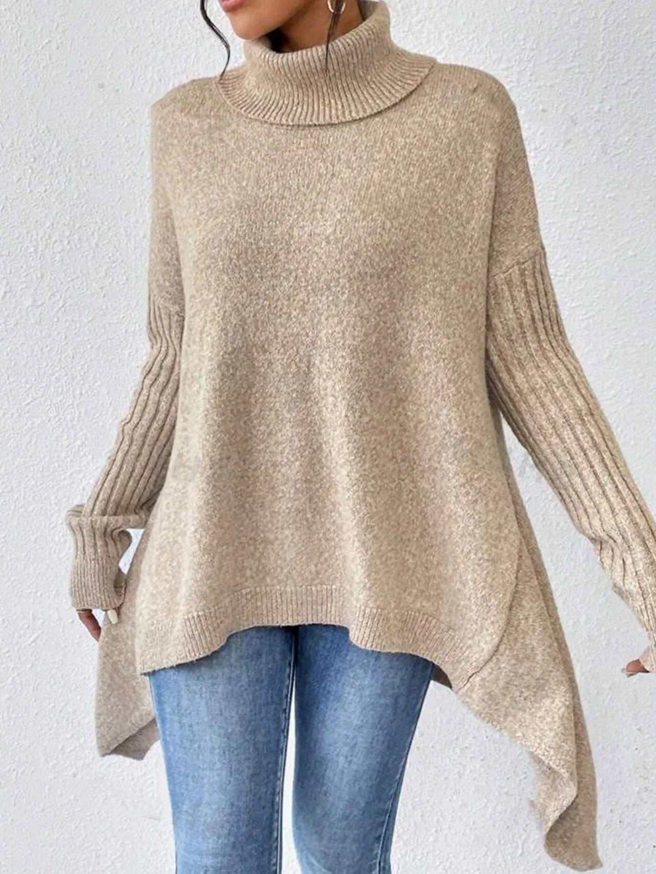Front view of a woman wearing a beige oversized turtleneck sweater with an asymmetrical hem and ribbed sleeves.