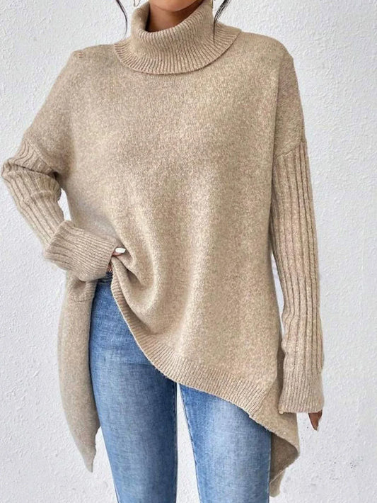 Woman wearing a beige oversized turtleneck sweater with an asymmetrical hem and ribbed sleeves, paired with blue jeans.