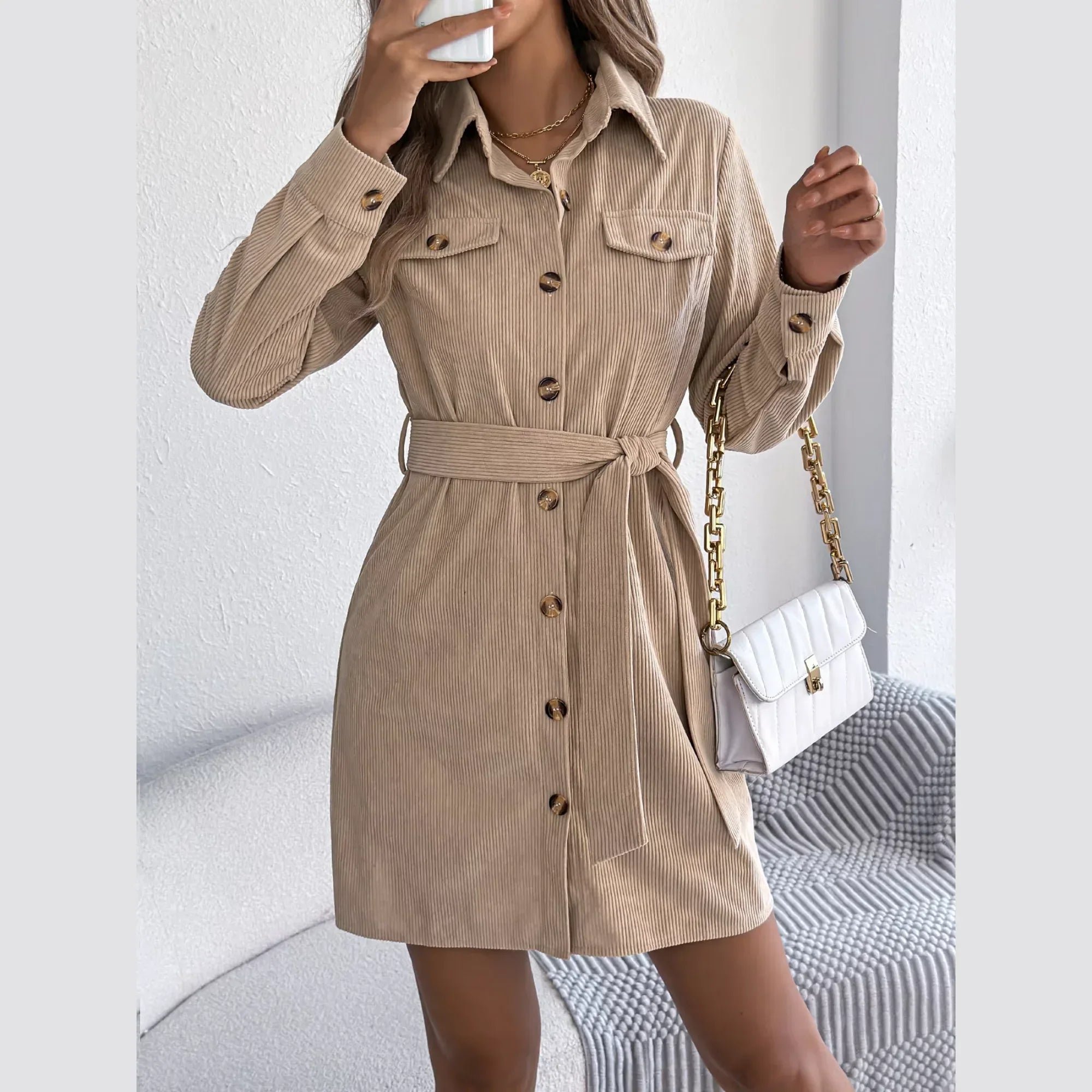 Woman holding a white quilted handbag and a phone while wearing a tan corduroy shirt dress.