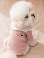 A white Bichon Frise dog wearing a pink ribbed knit sweater with a white ruffled collar and a small embroidered bunny motif on the chest.