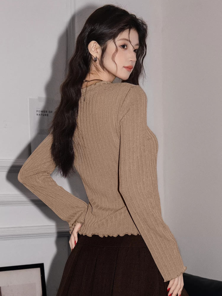 Back view of a woman wearing a tan ribbed knit long-sleeve top with a tie-front closure and ruffled hem, paired with a brown pleated skirt.
