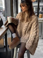 woman wearing a beige chunky cable knit oversized sweater with black tights and brown boots, holding a coffee cup