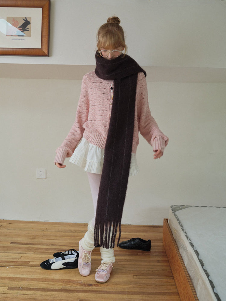 A young woman adjusts her dark scarf, which is draped over a pink knit cardigan and white pleated skirt. She is wearing white tights, leg warmers, and pink ballet flats.