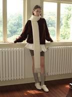Woman leaning on a window sill, wearing a brown velour sweatshirt with a white scarf, grey shorts, grey socks, and white sneakers.