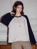 Close-up of a woman wearing a light grey long-sleeved t-shirt with navy blue raglan sleeves and beige pants, showing the embroidered detail.