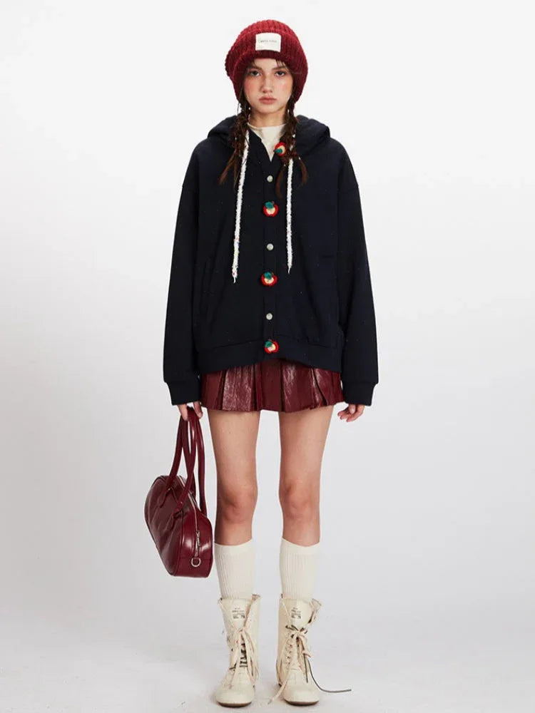 Woman wearing a dark blue hooded cardigan with colorful fruit buttons, a red beanie, a red faux leather pleated skirt, white knee-high socks, and cream-colored boots, holding a red handbag.