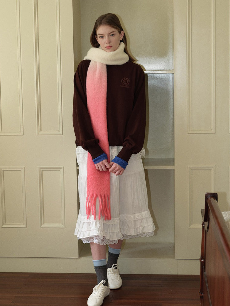 Full body shot of a woman wearing a brown sweatshirt, white ruffled skirt, and a long gradient scarf from cream to pink with fringe detail, standing.