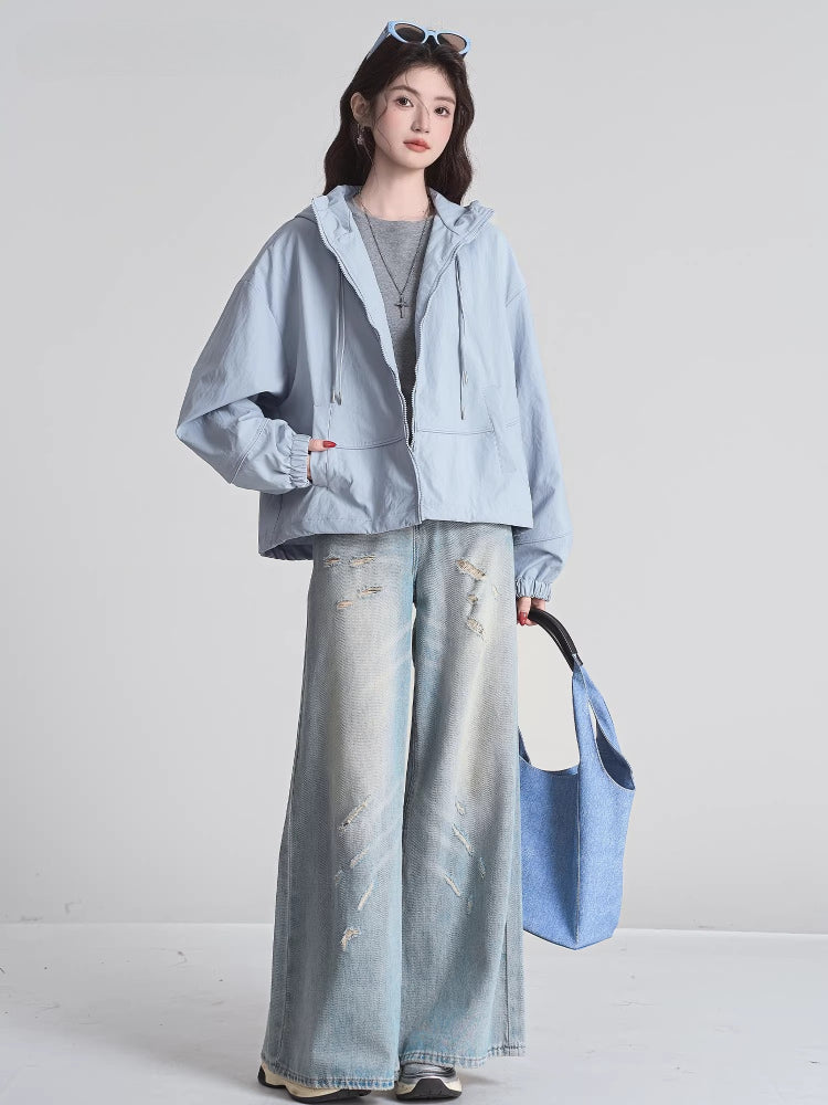 Woman wearing a light blue hooded jacket, grey t-shirt, and distressed wide-leg jeans, holding a blue tote bag.