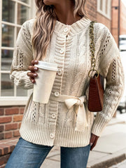 Woman wearing a cream cable knit cardigan tied with a belt, holding a white paper cup and a brown handbag.