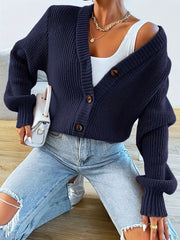 Woman kneeling on the floor wearing a navy blue oversized knit cardigan over a white tank top and ripped blue jeans, holding a white handbag and wearing white sneakers.