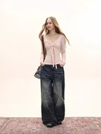 Full-body shot of a model wearing a light pink long-sleeve tie-front top and dark wash wide-leg jeans, standing on a patterned rug.