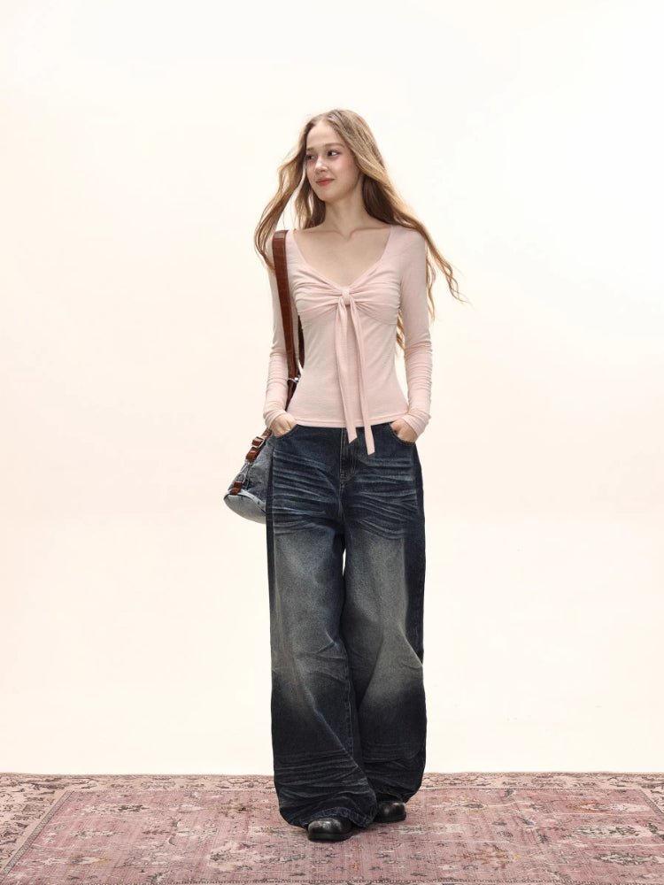 Full-body shot of a model wearing a light pink long-sleeve tie-front top and dark wash wide-leg jeans, standing on a patterned rug.