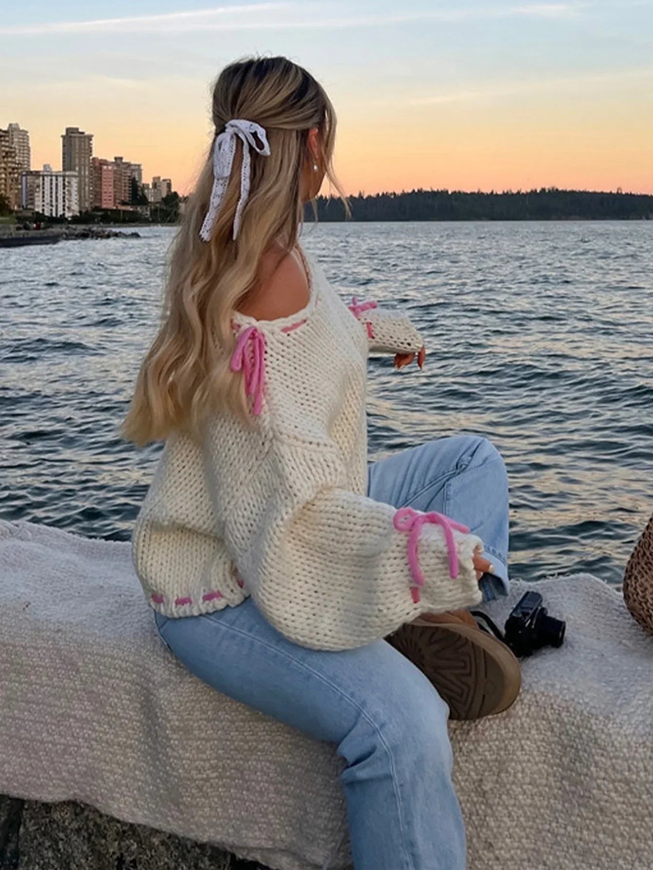 Woman with blonde hair wearing a cream chunky knit sweater with pink tie details, sitting by the water at sunset.