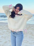 Woman with dark hair wearing a cream chunky knit sweater with pink stitching, posing on a beach.
