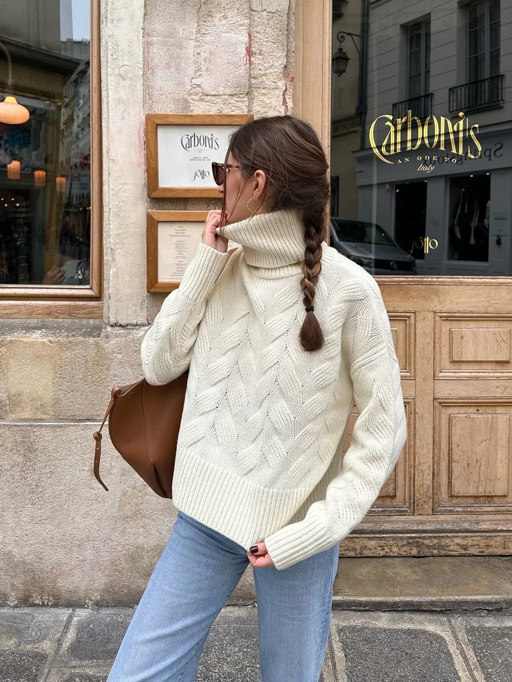 Woman in a cream cable knit turtleneck sweater and light wash jeans, adjusting her sunglasses.