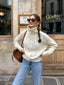 Woman wearing a cream cable knit turtleneck sweater with light wash jeans and a brown crossbody bag, standing in front of a building.