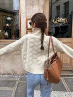 Back view of a woman wearing a cream cable knit turtleneck sweater with light wash jeans and a brown tote bag, with her arms outstretched.