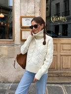 Woman in a cream cable knit turtleneck sweater and light wash jeans, looking over her shoulder.