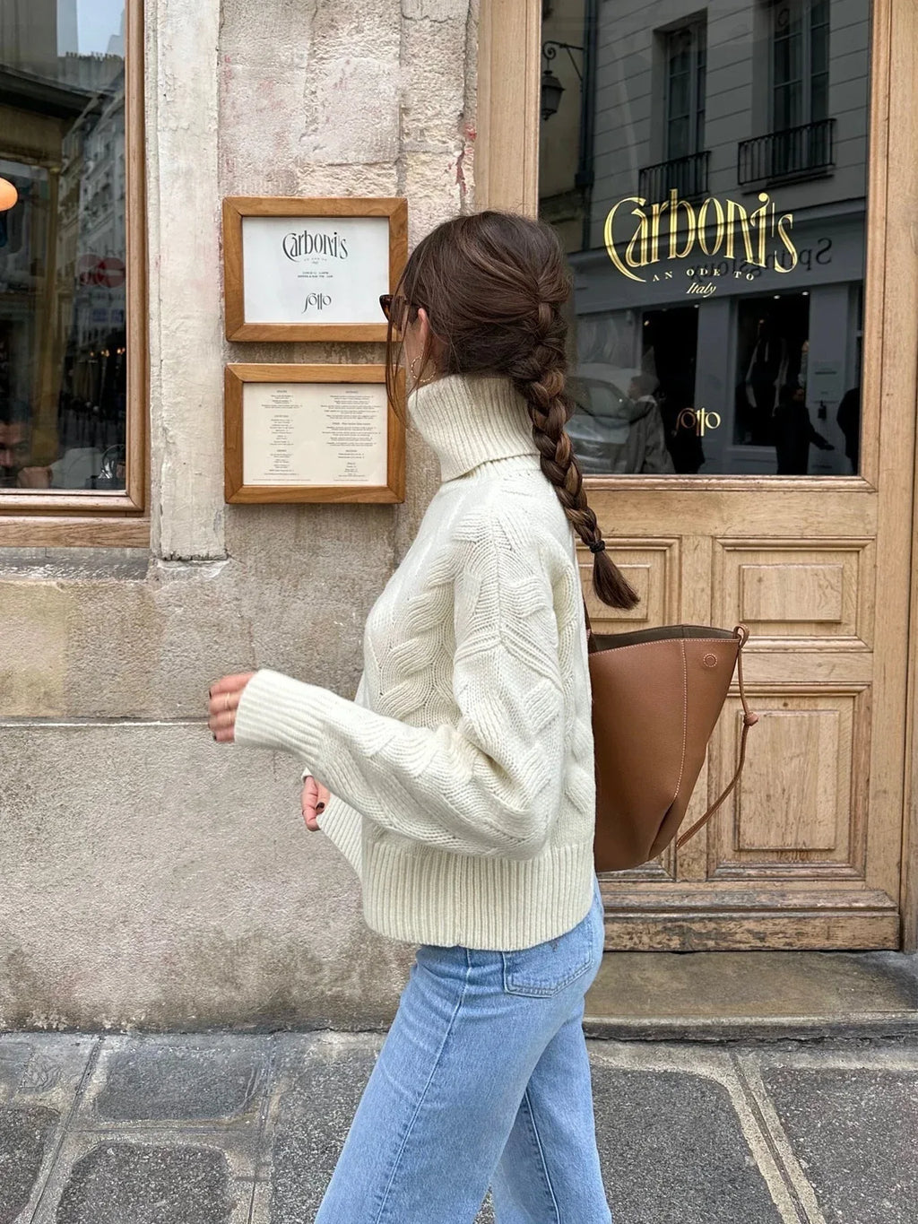 Side view of a woman wearing a cream cable knit turtleneck sweater and light wash jeans, walking.