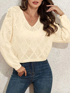 Front view of a woman wearing a cream-colored V-neck cropped knit sweater with long sleeves and a diamond pattern, styled with blue jeans and a delicate necklace.