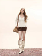 Full-body shot of a model in a cream long-sleeve top with lace trim and a bow detail, brown shorts, and cream slouchy boots.