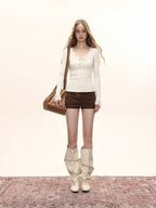 Model standing with hands in pockets, wearing a cream long-sleeve top with lace trim and a bow detail, and brown shorts.