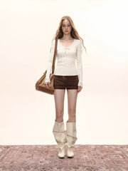 Model standing with hands in pockets, wearing a cream long-sleeve top with lace trim and a bow detail, and brown shorts.