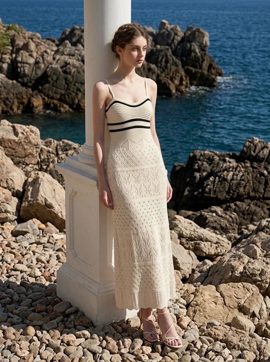 Woman wearing a cream knit maxi dress with black stripes on the bust, standing next to a pillar on a rocky coastline.