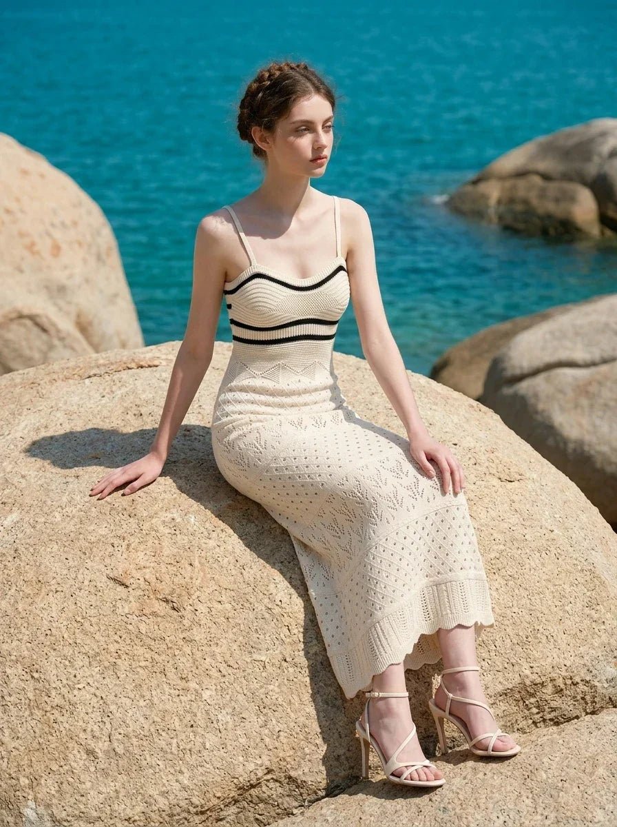 Woman wearing a cream knit maxi dress with black stripes on the bust, sitting on a large rock by the ocean, looking out to sea.