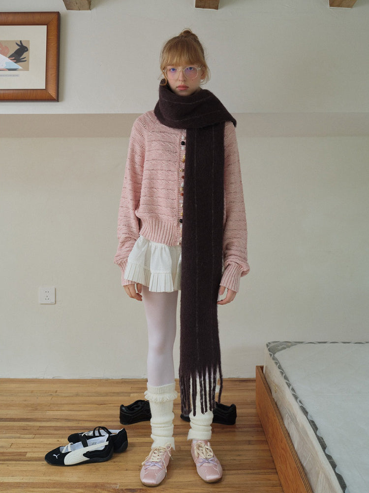 A young woman stands in a room wearing a pink knit cardigan over a white pleated skirt, a long dark scarf, white tights, leg warmers, and pink ballet flats. Black and white sneakers are on the floor.