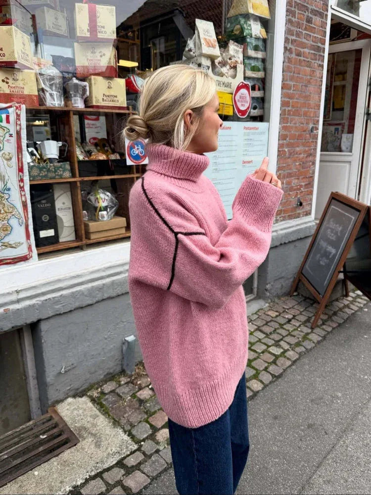 Woman wearing a pink oversized turtleneck sweater with contrasting black seam details and dark wash jeans, standing outside a shop.