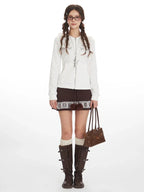 Woman wearing a white zip-up hoodie, a brown patterned skirt, white socks, and brown boots, holding a brown handbag.