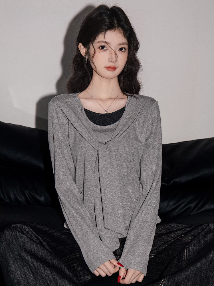 Woman wearing a grey long-sleeved top with a draped overlay and black pants, sitting on a black couch.