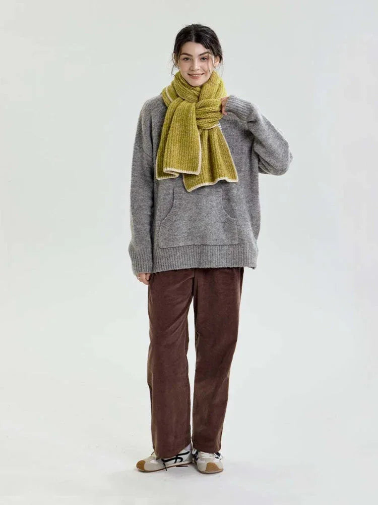 Model standing, showcasing a lime green chunky knit scarf styled over a grey sweater and brown corduroy pants.