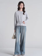 Full body shot of a woman wearing a light grey knit cardigan with a drawstring waist and button front closure, paired with blue jeans and a white handbag.